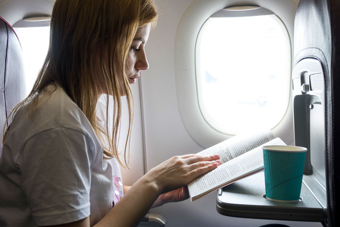 Passenger reading a book by the airplane window with a cup on the tray table during a flight. Passenger reading a book by the airplane window with a cup on the tray table during a flight.