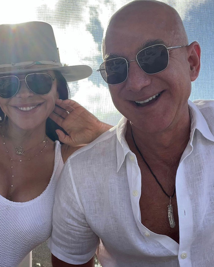Jeff Bezos and Lauren Sanchez smiling together outdoors wearing sunglasses and casual white outfits. Jeff Bezos and Lauren Sanchez smiling together outdoors wearing sunglasses and casual white outfits.