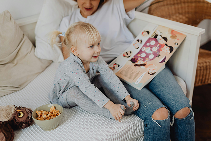 Young woman and child stretching on a bed with a book and snacks, reflecting family dynamics and relationship tensions. Young woman and child stretching on a bed with a book and snacks, reflecting family dynamics and relationship tensions.