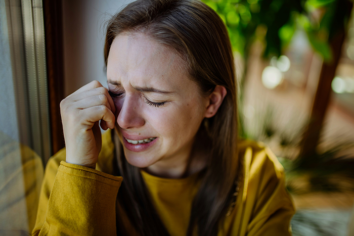 Young woman in a mustard sweater crying indoors near window, expressing sorrow on a ghost friend birthday memory.