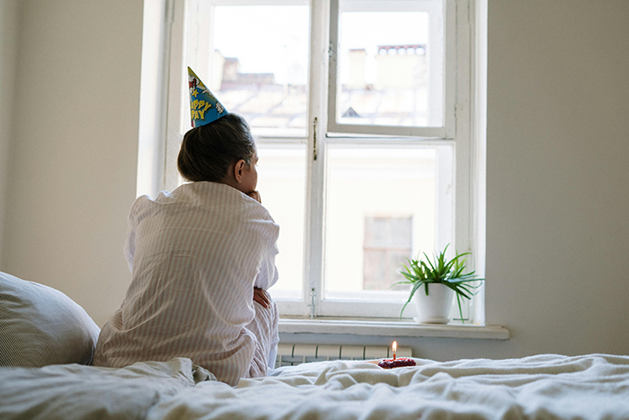 Person wearing a birthday hat sitting alone by a window with a small lit cake, reflecting on longtime friends and ghost friend birthday.