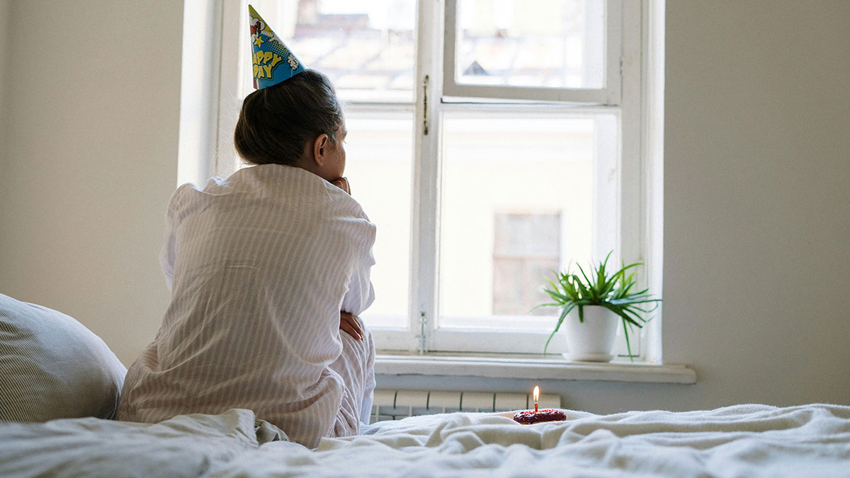 Woman in birthday hat sitting alone on bed with single lit candle, reflecting on ladyu2019s themed 30th bday party ruined.