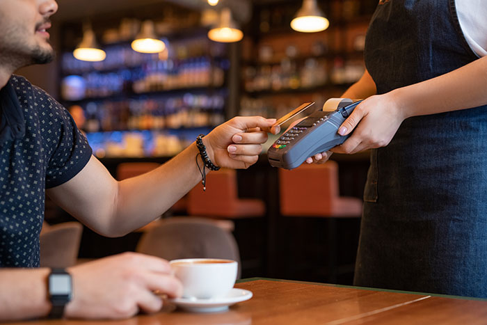 Customer tipping 83 cents while paying with a card in a cafe, barista holding a payment terminal in a casual setting. Customer tipping 83 cents while paying with a card in a cafe, barista holding a payment terminal in a casual setting.