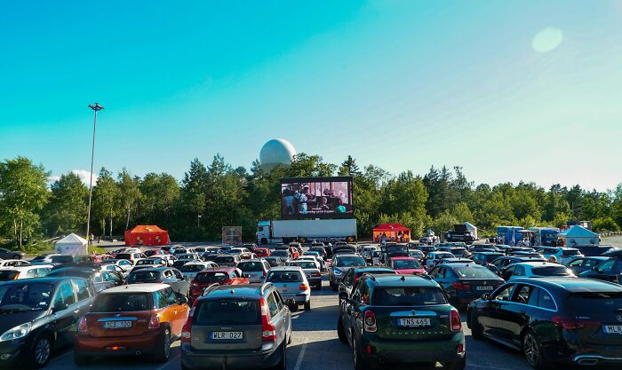 Cars parked at an outdoor drive-in movie theater under a clear blue sky with trees in the background.