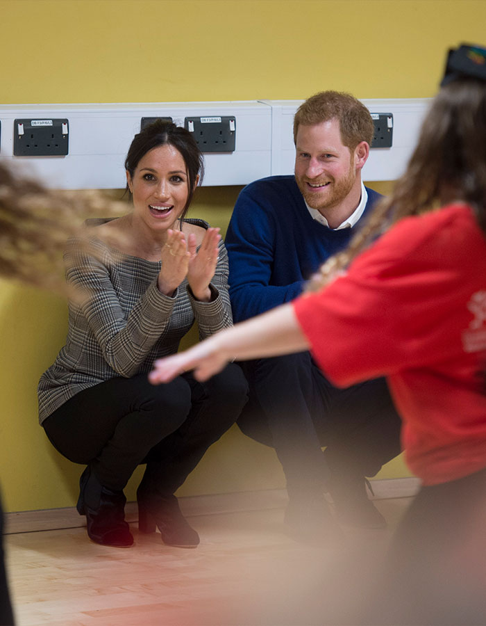 Meghan Markle and Prince Harry smiling and clapping while watching a dance session in a room with yellow walls. Meghan Markle and Prince Harry smiling and clapping while watching a dance session in a room with yellow walls.