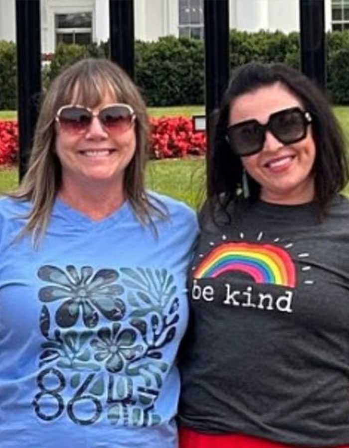 Two women wearing sunglasses and graphic t-shirts, standing outside near the White House with flowers in the background. Two women wearing sunglasses and graphic t-shirts, standing outside near the White House with flowers in the background.