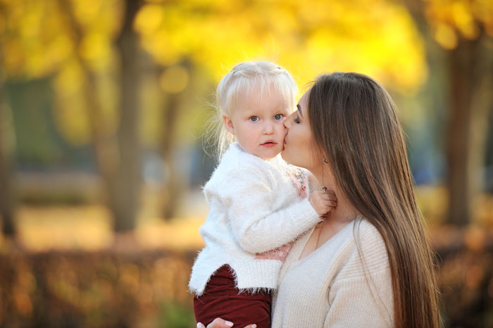Mother kissing her mil daughter outdoors during a surprise vacation in an autumn park with golden leaves.