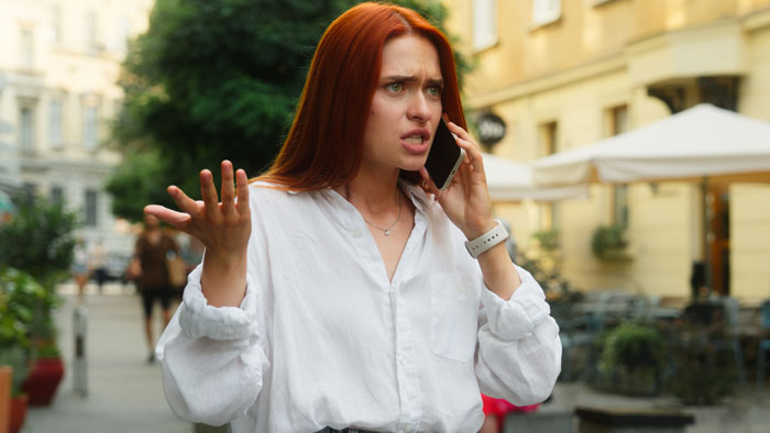 Red-haired woman on phone looking upset outdoors, expressing frustration about mil-daughter-surprise-vacation plans.