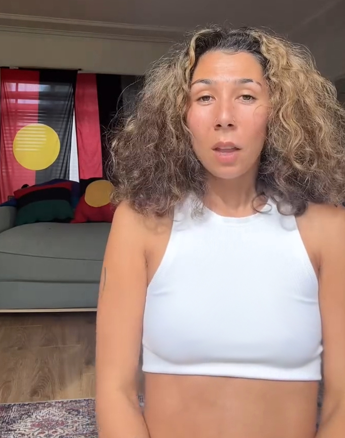 Aboriginal artist with curly hair in white top, speaking indoors with Aboriginal flag and cushions in background. Aboriginal artist with curly hair in white top, speaking indoors with Aboriginal flag and cushions in background.