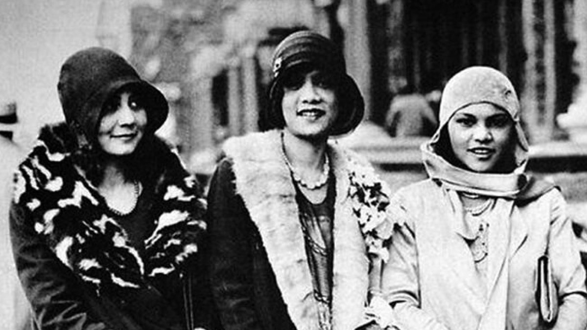 Three women dressed in Harlem Renaissance fashion, wearing cloche hats and fur-trimmed coats on a city street.