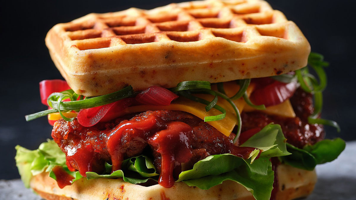 Close-up of a mouthwatering waffle sandwich with fried chicken, lettuce, cheese, and sauce in vibrant food photography.