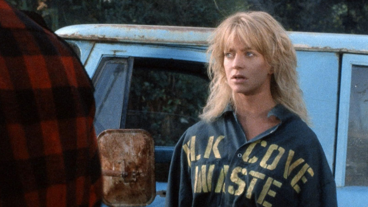 Blonde woman in a dark shirt with yellow letters standing near a blue vehicle, illustrating toxic romantic movies theme.
