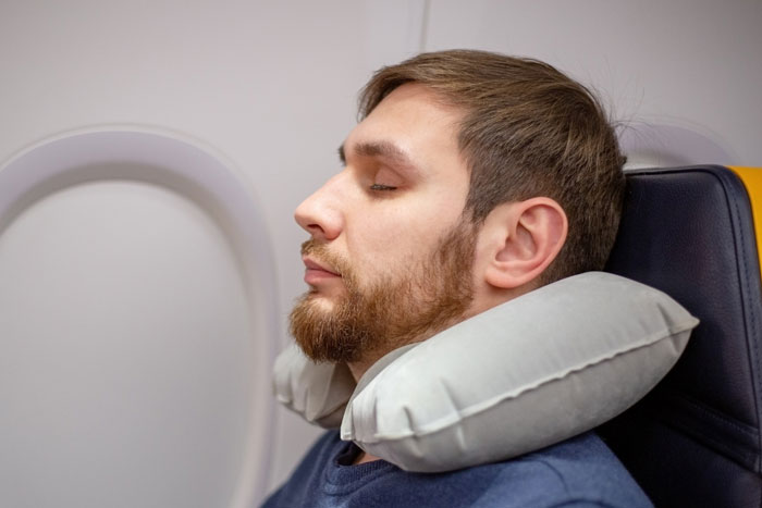 Passenger napping on airplane seat with headrest pillow, unaware of obnoxious guy about to slap his head.