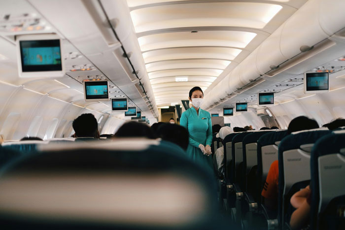 Flight attendant in teal uniform walks down airplane aisle among passengers, while obnoxious guy slaps napping passenger’s head incident unfolds.
