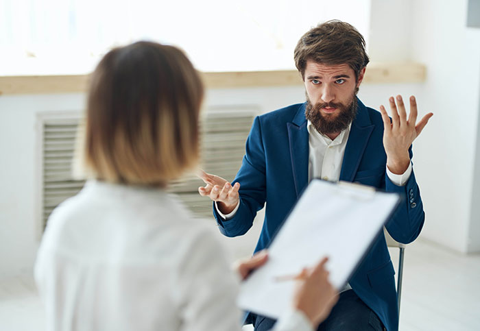 Man in blue suit explaining while woman with clipboard listens in a workplace, non-profit employee hour dispute discussion. Man in blue suit explaining while woman with clipboard listens in a workplace, non-profit employee hour dispute discussion.