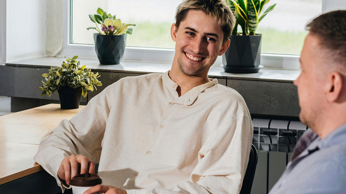 Two coworkers smiling and chatting inside office near window with plants, illustrating not in job description petty revenge concept.