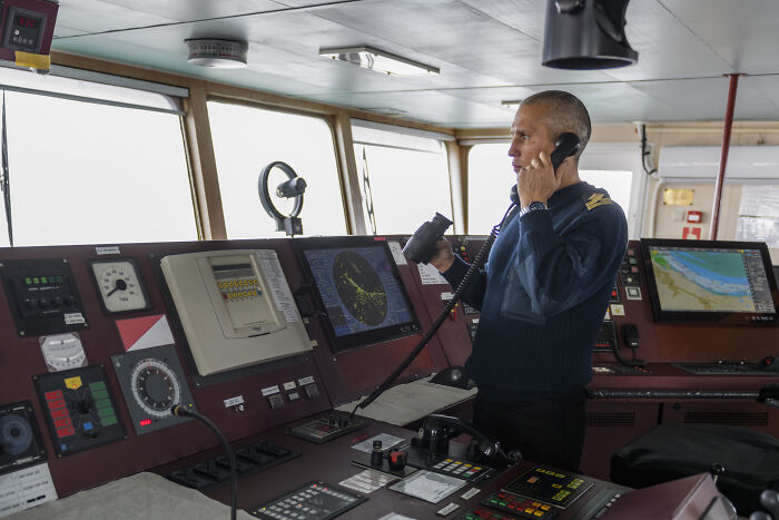 Man in a navy uniform using a radio on a ship’s bridge surrounded by navigation and communication equipment, representing professions.