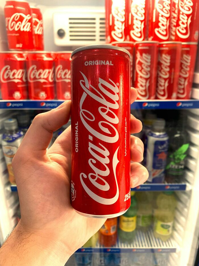 Hand holding a Coca-Cola can in front of a fridge stocked with Coke cans and various beverages.