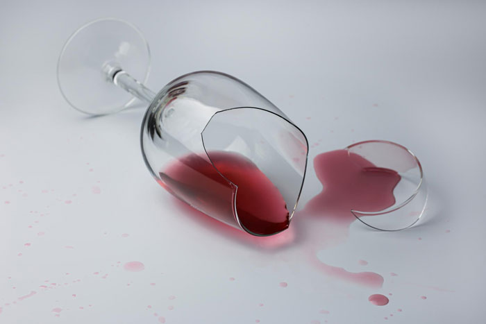 Broken wine glass spilled with red liquid on white surface symbolizing damages to dishes and furniture in a relationship conflict. Broken wine glass spilled with red liquid on white surface symbolizing damages to dishes and furniture in a relationship conflict.