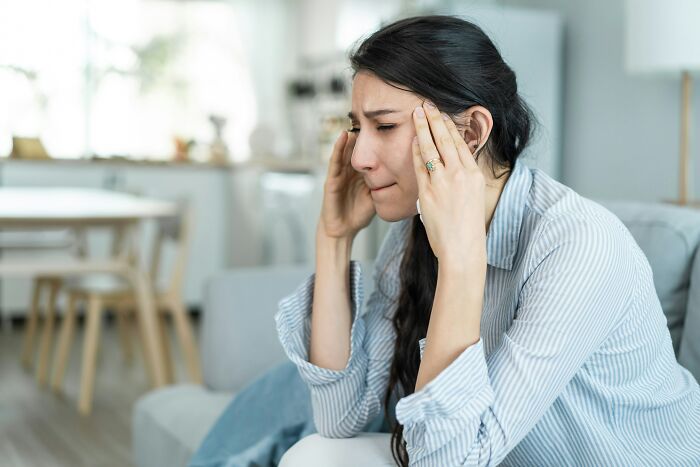Stressed woman sitting on couch, holding temples, showing signs of insecurity and emotional distress indoors.