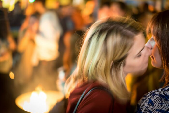 Two women sharing a kiss at a crowded social event, capturing a moment of intimacy amid blurred background activity.