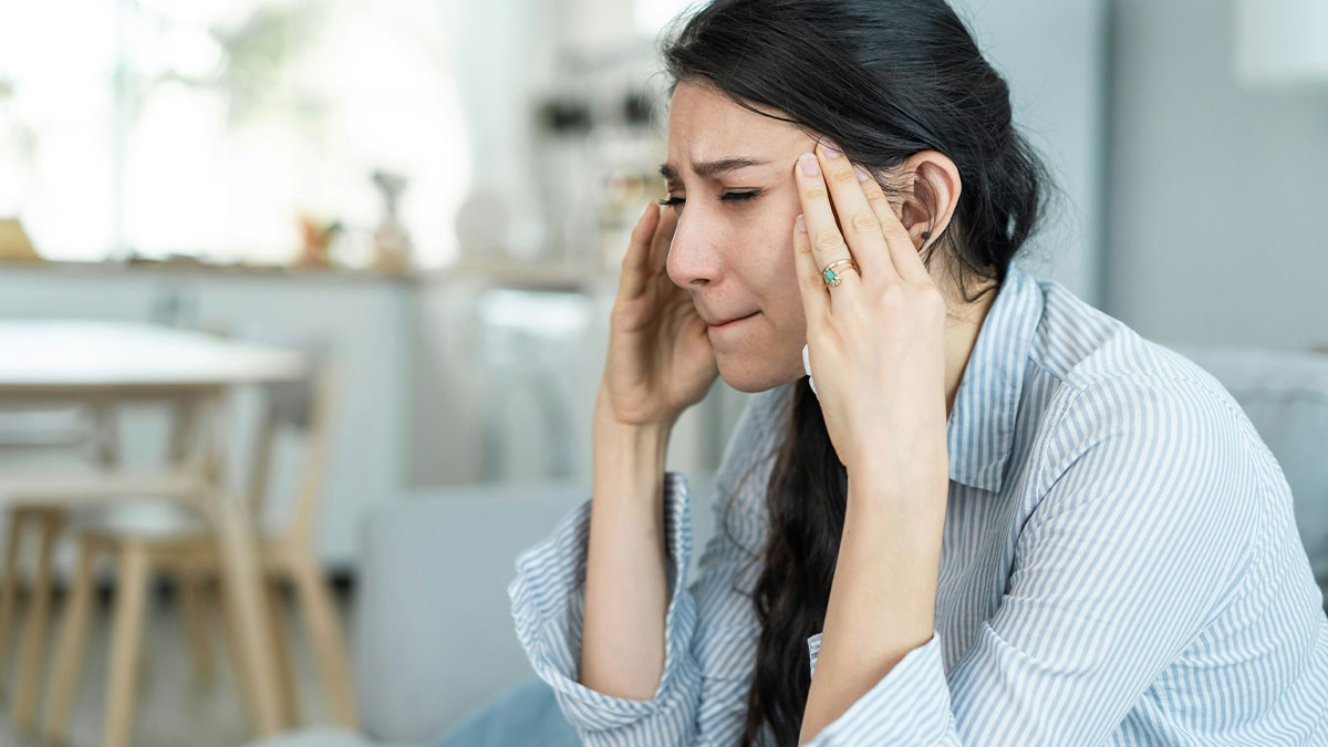 Stressed woman sitting indoors, holding temples with eyes closed, showing signs of insecurity and emotional distress.