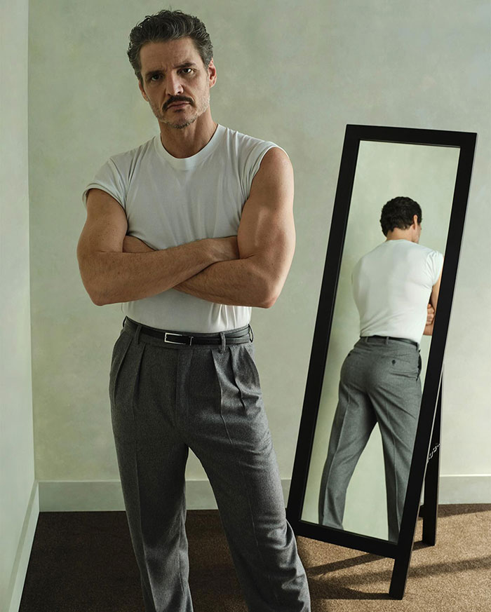 Pedro Pascal standing with arms crossed wearing a white shirt and gray pants next to a full-length mirror. Pedro Pascal standing with arms crossed wearing a white shirt and gray pants next to a full-length mirror.