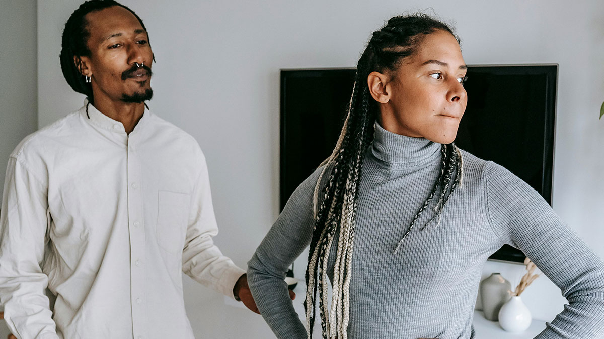 A woman looks away with crossed arms while a man tries to talk to her, showing subtle signs of low self-esteem.