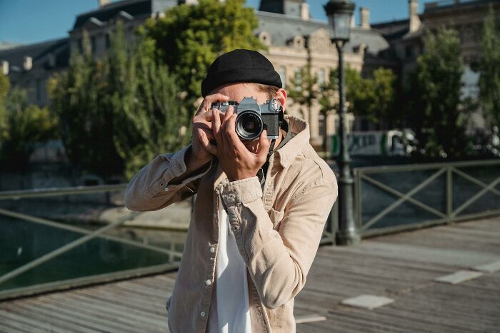 Person wearing a beige jacket and black cap taking a photo outdoors, illustrating subtle signs of low self-esteem.
