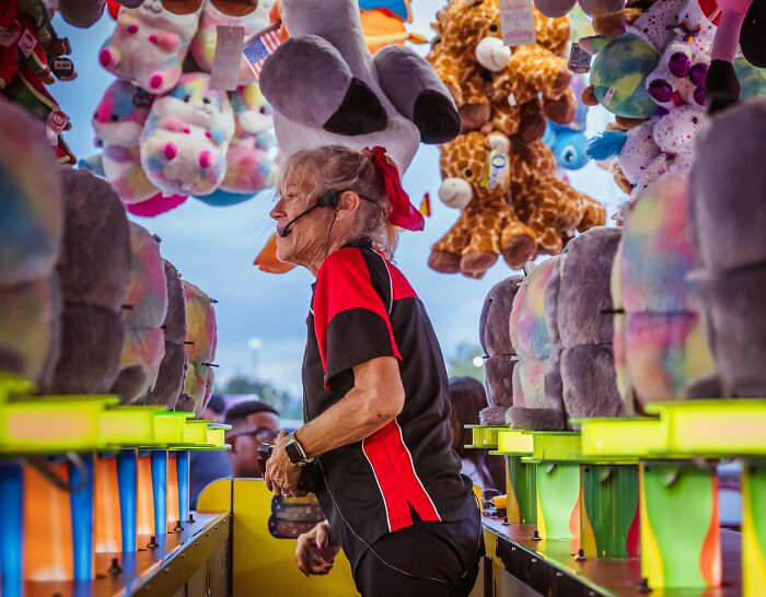 Woman in headset working at a carnival game booth surrounded by stuffed animal prizes, highlighting professions attracting awful people