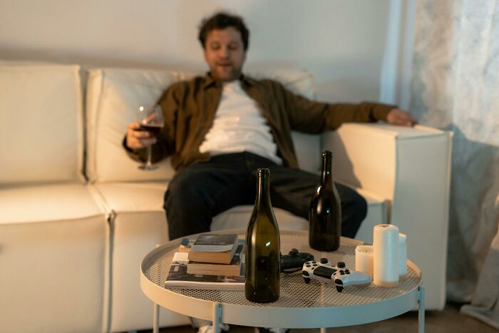 Man sitting on a couch with wine glass, surrounded by bottles and gaming controllers, depicting life ruined by one wrong deed.