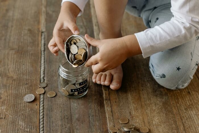 Child in pajamas pouring coins into a glass jar on wooden floor, representing relatable middle class money habits.