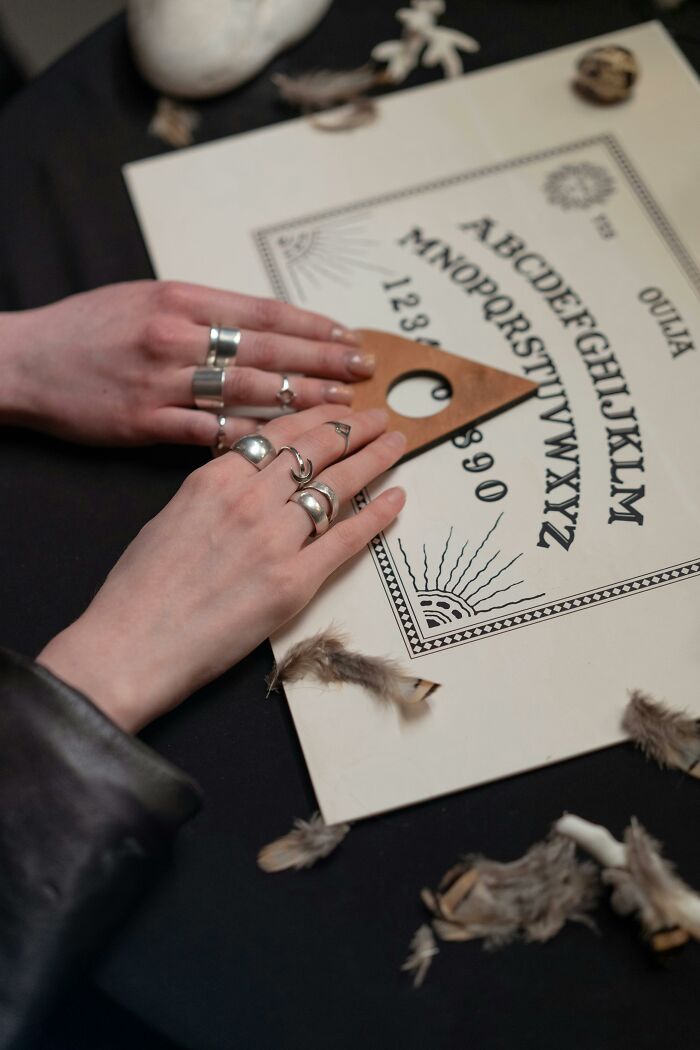 Hands with silver rings using a wooden planchette on a Ouija board surrounded by feathers about afterlife theories.