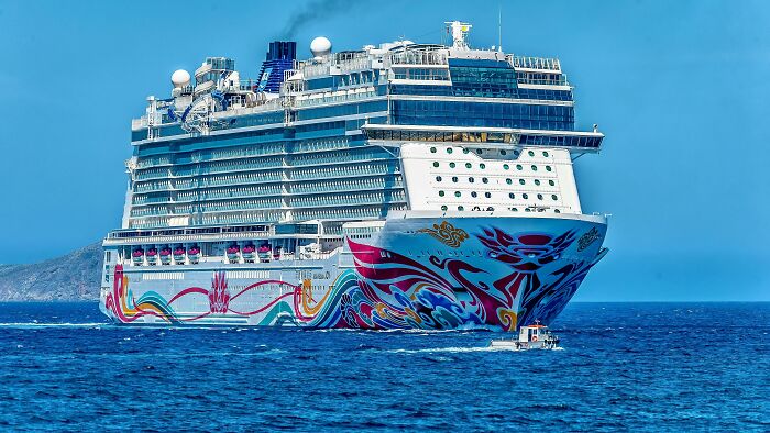 A large cruise ship with colorful flags and patterns on its hull sailing on calm blue ocean waters under a clear sky.