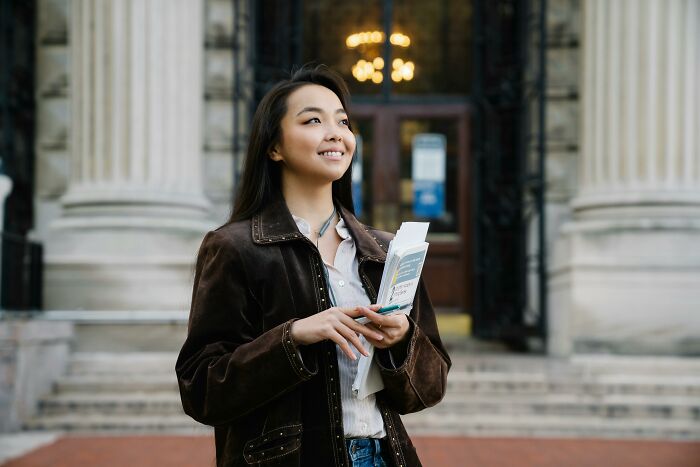 Young adult smiling outside a school building, holding papers, reflecting adults who never moved on from their glory days in high school