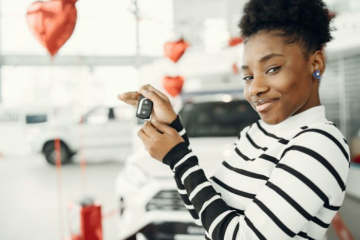 Young woman holding car keys at dealership smiling proudly, relatable middle class moment captured indoors with vehicles and balloons.