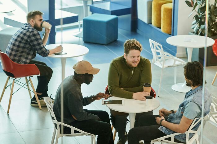 Three men sharing privileged things said while having coffee in a modern cafe setting with another man drinking alone nearby.