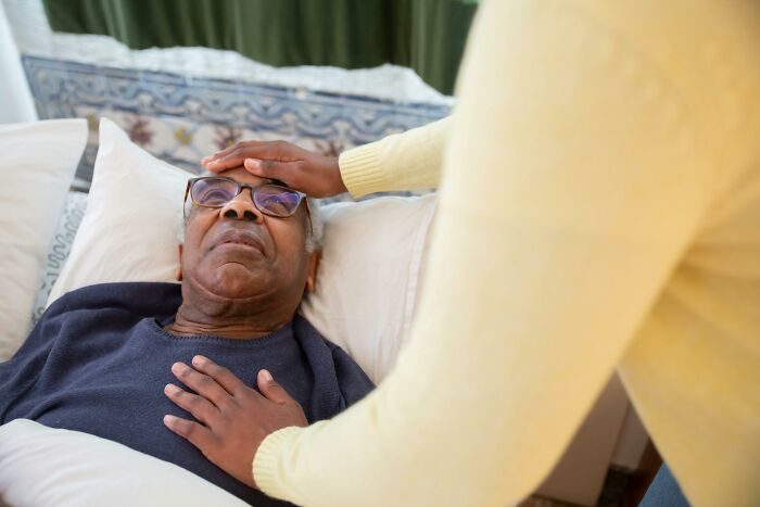 Elderly man lying in bed with a caregiver checking his forehead, illustrating reporting coworker diabolical things to HR.