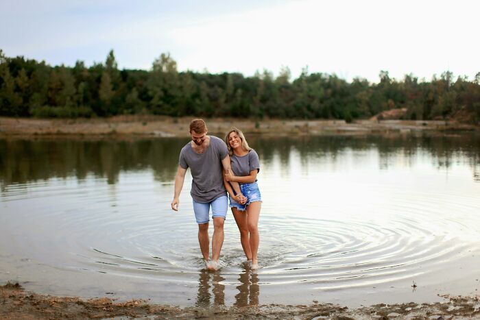 Couple reliving high school glory days, walking barefoot in a lake near a forest on a calm day.