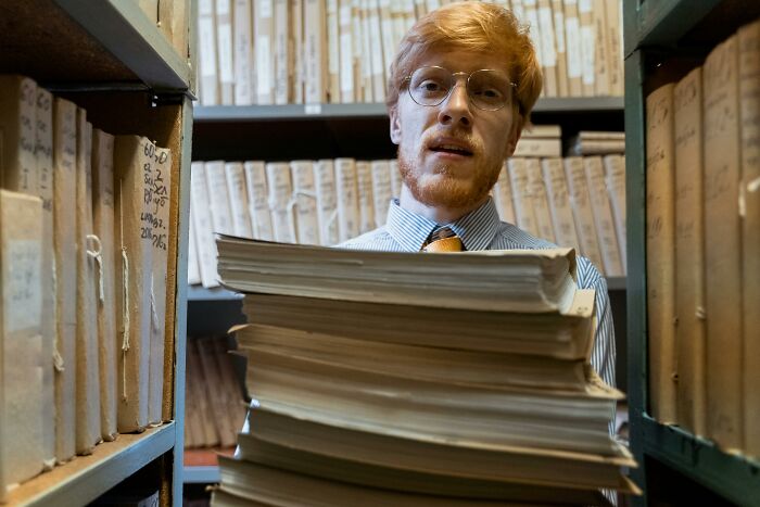 Man wearing glasses carrying large stack of documents in archive room, representing ordering sequences challenge. Man wearing glasses carrying large stack of documents in archive room, representing ordering sequences challenge.