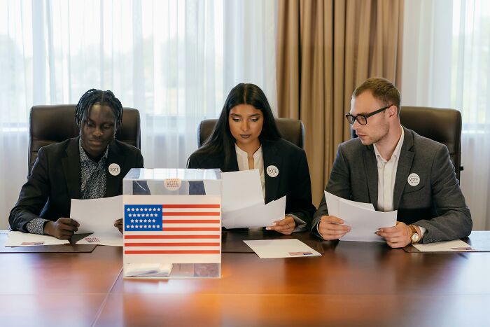 Three people wearing vote badges examining papers at a table with a transparent ballot box showing a US flag, reflecting a good trend slowly disappeared.