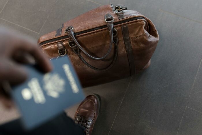 Traveler holding passport near brown leather bag on airport floor to avoid TSA security line delay. Traveler holding passport near brown leather bag on airport floor to avoid TSA security line delay.
