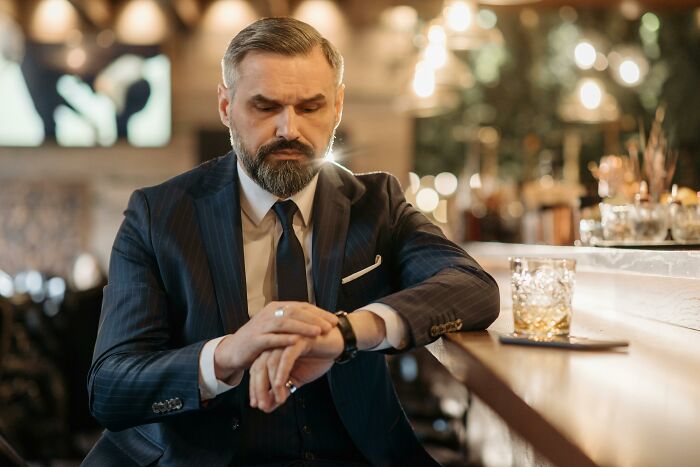 Middle-aged man in a suit looking at his watch, sitting at a bar with a glass of whiskey nearby, reflecting on sour dates.