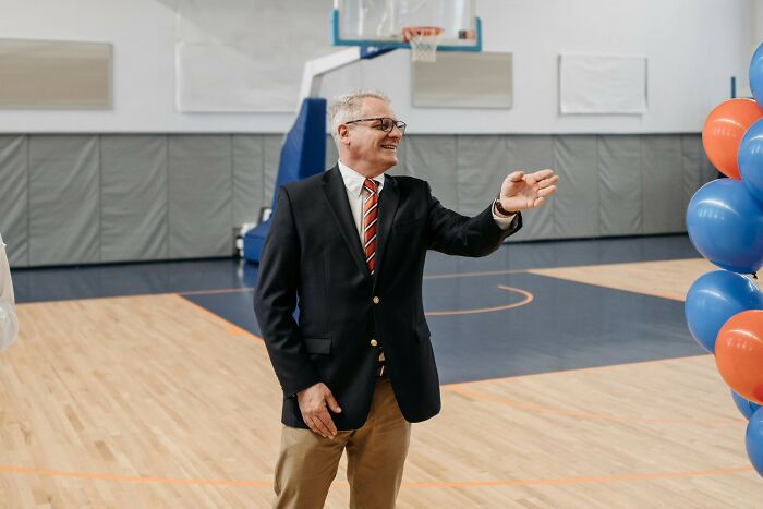Man in a suit gestures and smiles in a gymnasium decorated with blue and orange balloons, representing professions attracting awful people.
