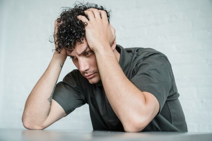 Young man with curly hair holding his head, showing subtle signs of incredibly low self-esteem in a casual setting.