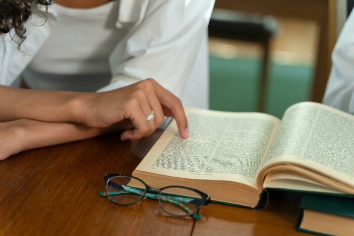 Person pointing at an open book on a wooden table with eyeglasses nearby, related to vocabulary challenge words. Person pointing at an open book on a wooden table with eyeglasses nearby, related to vocabulary challenge words.