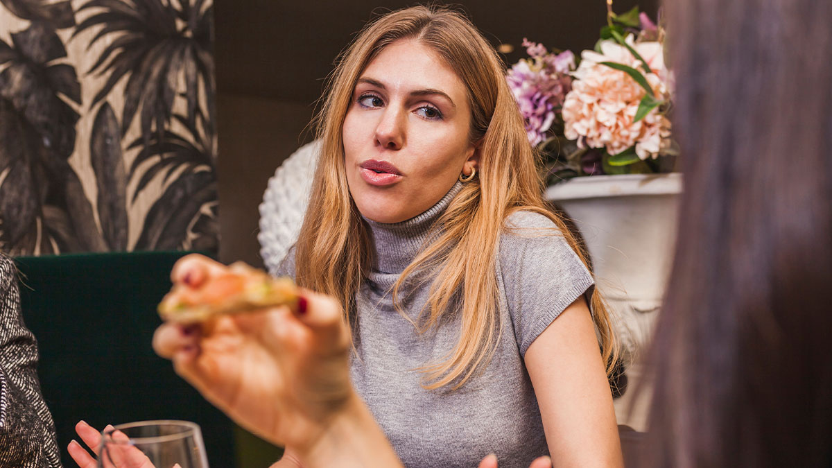 Woman having a serious conversation while sitting indoors, showing emotion in a social setting with flowers in background.