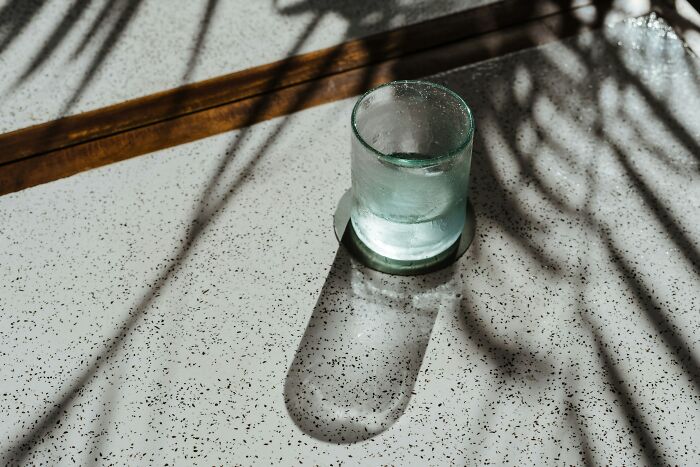 Glass of water casting shadow on speckled table under natural light with plant shadows in a calm setting.