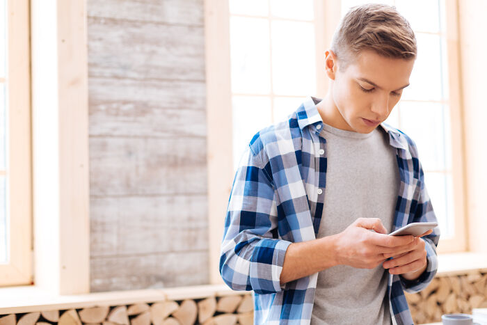 Teen boy in a plaid shirt looking at phone, illustrating rich kid meltdowns at private schools in a bright room.
