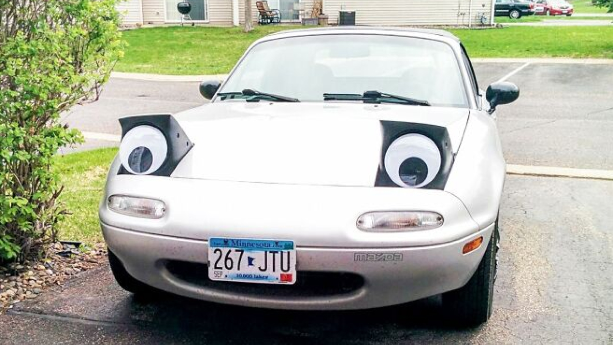 Silver Mazda car with quirky large googly eyes attached to its headlights in a suburban driveway setting.